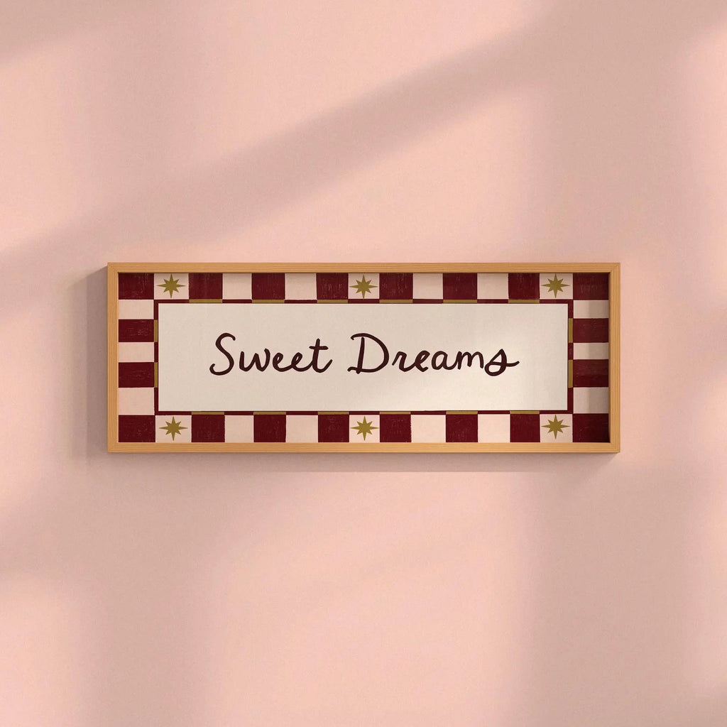 A rectangular framed wall sign with the words 'Sweet Dreams' in dark brown script, set against a cream center panel and a red-and-cream checkered border with small gold star accents, mounted on a soft pink wall.