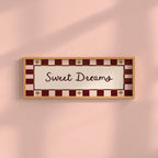 A rectangular framed wall sign with the words 'Sweet Dreams' in dark brown script, set against a cream center panel and a red-and-cream checkered border with small gold star accents, mounted on a soft pink wall.