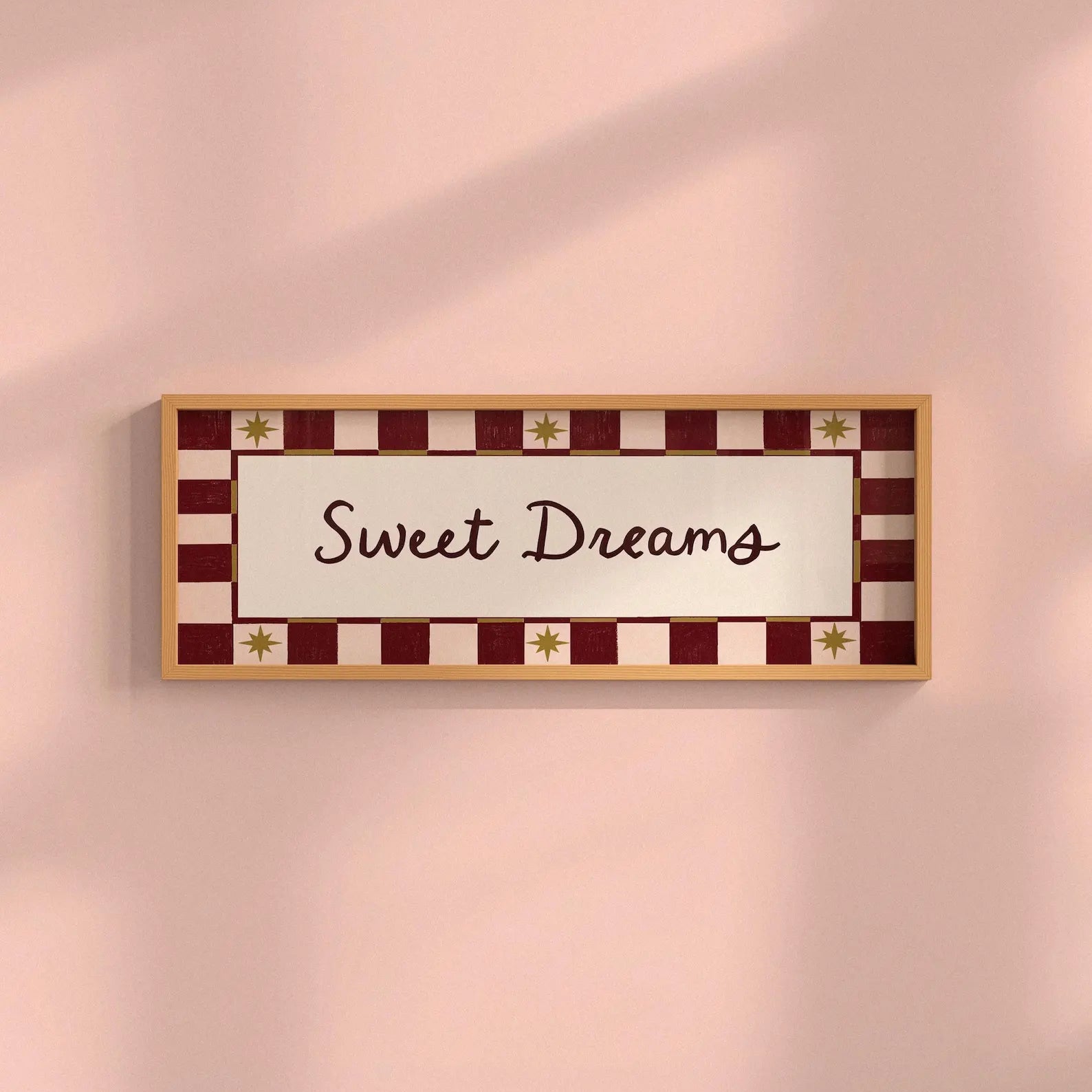 A rectangular framed wall sign with the words 'Sweet Dreams' in dark brown script, set against a cream center panel and a red-and-cream checkered border with small gold star accents, mounted on a soft pink wall.