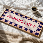 A rectangular novelty sign with the words 'DRAMATIC EXIT' and an arrow, styled on white bedding with a cup of coffee and eyeglasses nearby.