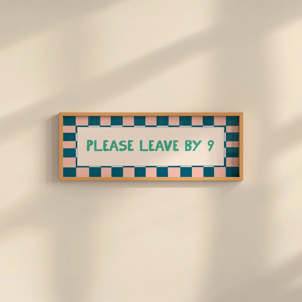 A rectangular framed sign on a warm beige wall with soft diagonal shadows. The sign has a light wood frame, a white center, teal text reading 'PLEASE LEAVE BY 9', and a border of alternating teal and blush pink squares.