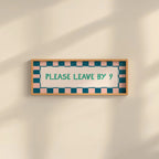 A rectangular framed sign on a warm beige wall with soft diagonal shadows. The sign has a light wood frame, a white center, teal text reading 'PLEASE LEAVE BY 9', and a border of alternating teal and blush pink squares.