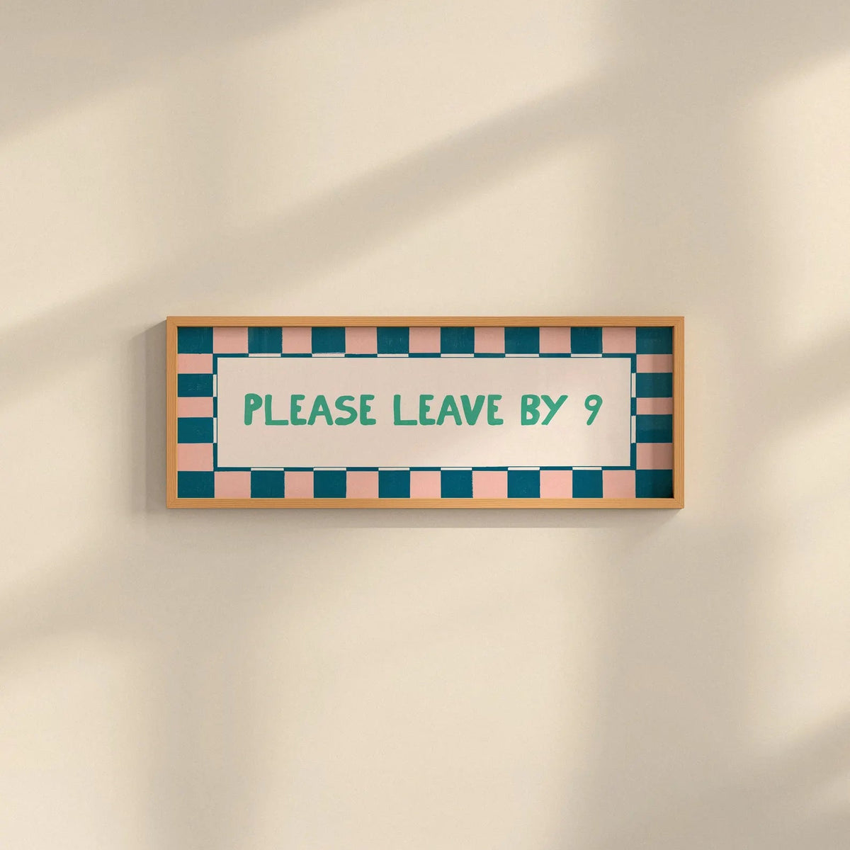 A rectangular framed sign on a warm beige wall with soft diagonal shadows. The sign has a light wood frame, a white center, teal text reading 'PLEASE LEAVE BY 9', and a border of alternating teal and blush pink squares.