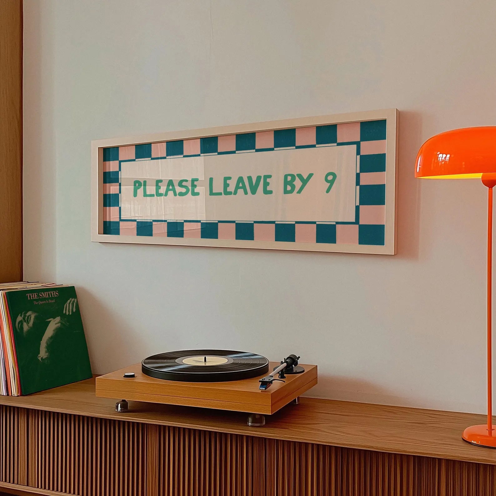 A framed wall sign with a pink and teal checkered border and the text 'PLEASE LEAVE BY 9' in teal, displayed above a wooden record player on a cabinet in a styled room setting.