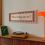 A framed wall art print with the words 'Love you, bye' in dark red script over a striped background, hanging on a light wall above a wooden credenza with a turntable and an orange floor lamp.