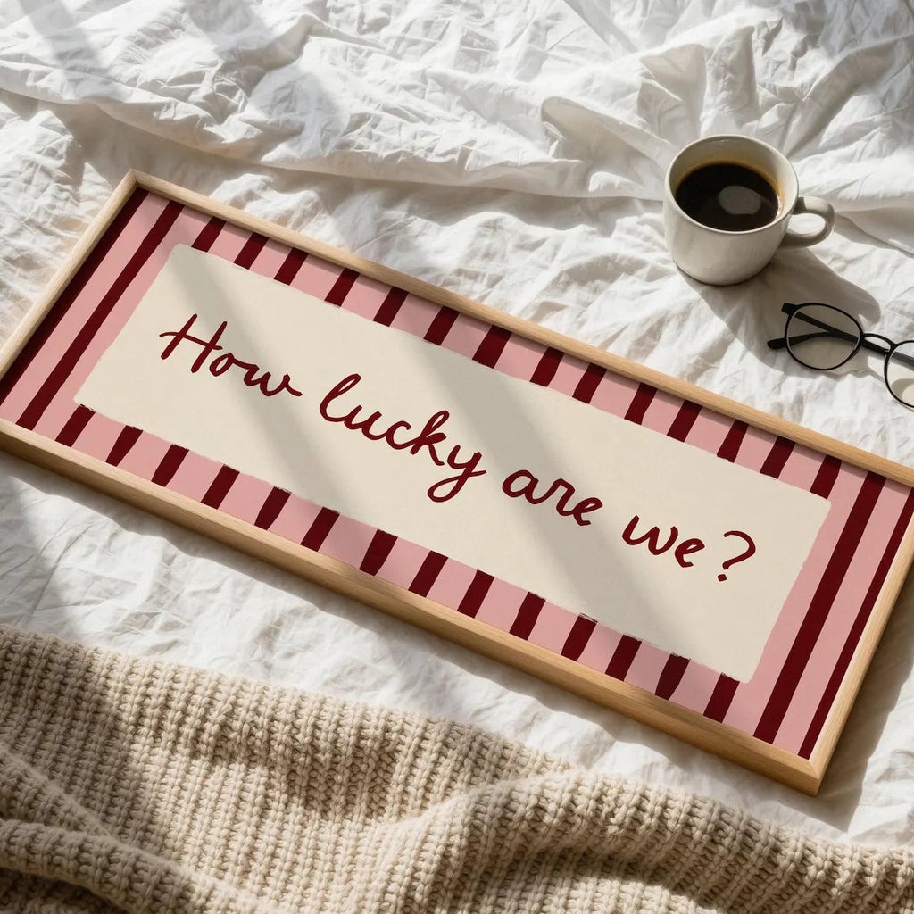 A framed decorative sign reading “How lucky are we?” in red script, placed on rumpled white bedding with a mug, glasses, and a knitted blanket nearby.