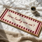 A framed decorative sign reading “How lucky are we?” in red script, placed on rumpled white bedding with a mug, glasses, and a knitted blanket nearby.