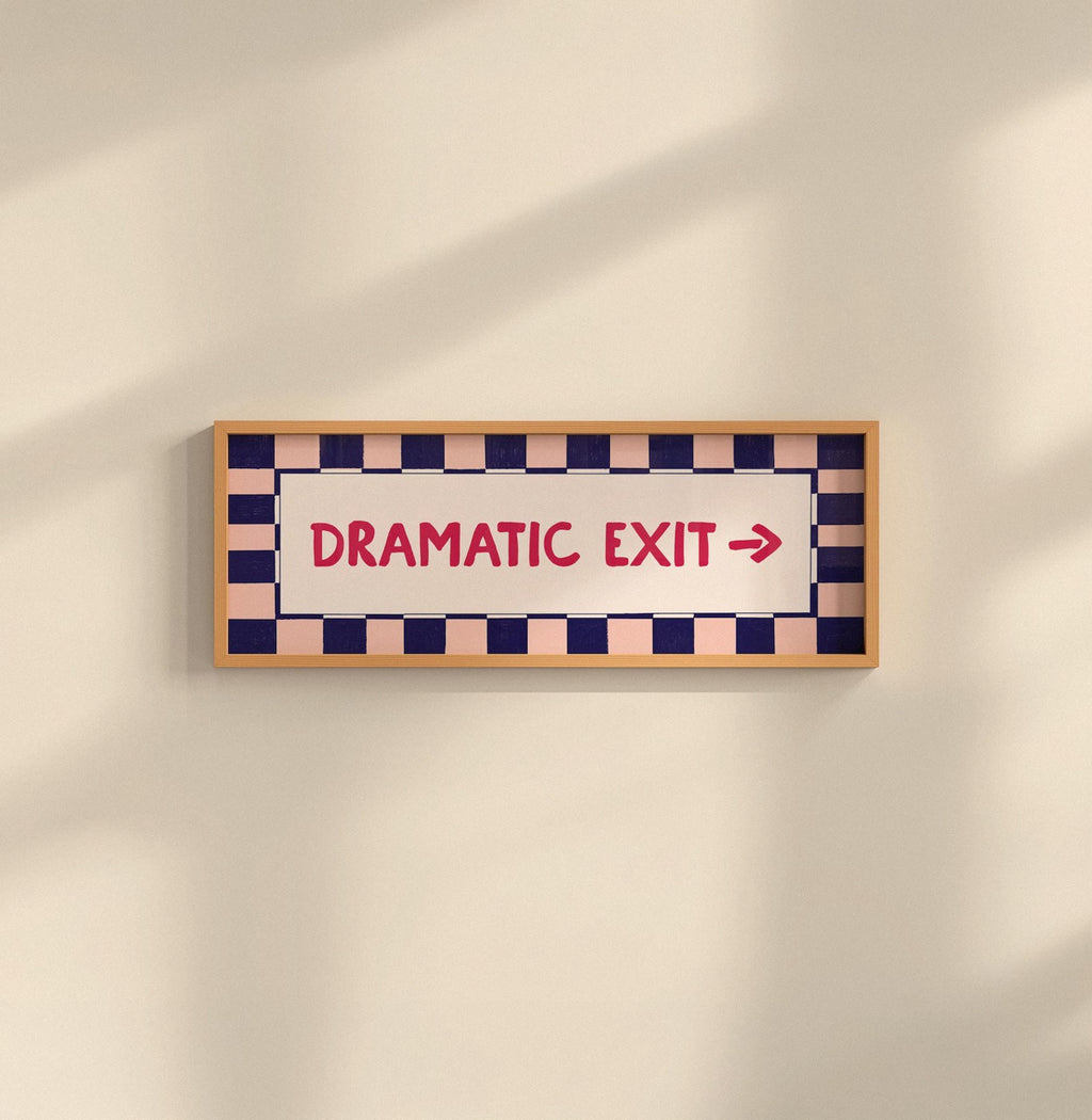 A rectangular framed sign on a beige wall with a wood frame, navy and peach checkered border, and red text reading 'DRAMATIC EXIT' with a right arrow.