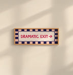 A rectangular framed sign on a beige wall with a wood frame, navy and peach checkered border, and red text reading 'DRAMATIC EXIT' with a right arrow.