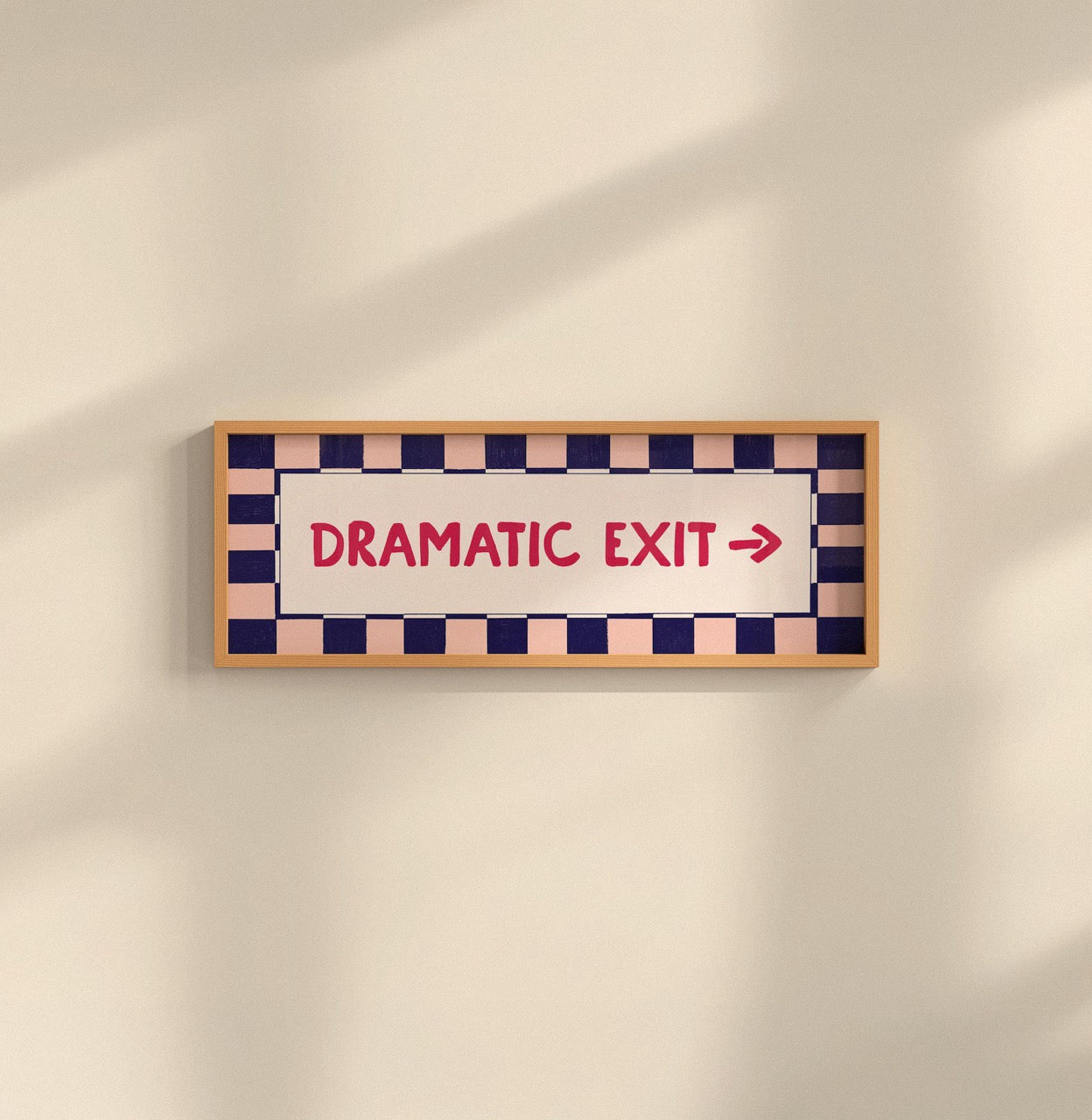 A rectangular framed sign on a beige wall with a wood frame, navy and peach checkered border, and red text reading 'DRAMATIC EXIT' with a right arrow.