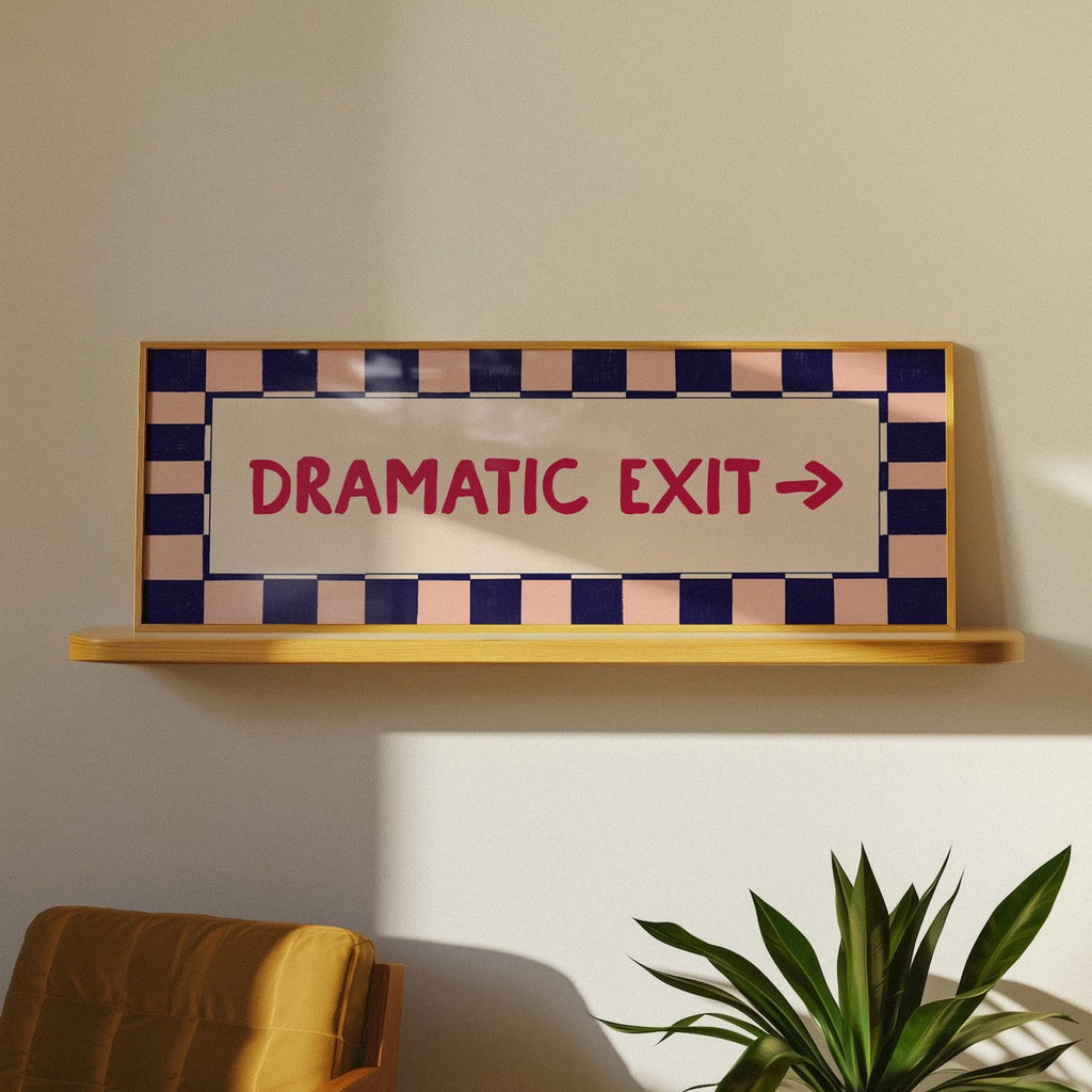A decorative emergency exit sign with the words 'DRAMATIC EXIT' and a right arrow, mounted on a light wood shelf against a beige wall. The sign has a cream center panel, red lettering, and a checkered border in navy blue and pale pink.