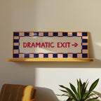 A decorative emergency exit sign with the words 'DRAMATIC EXIT' and a right arrow, mounted on a light wood shelf against a beige wall. The sign has a cream center panel, red lettering, and a checkered border in navy blue and pale pink.