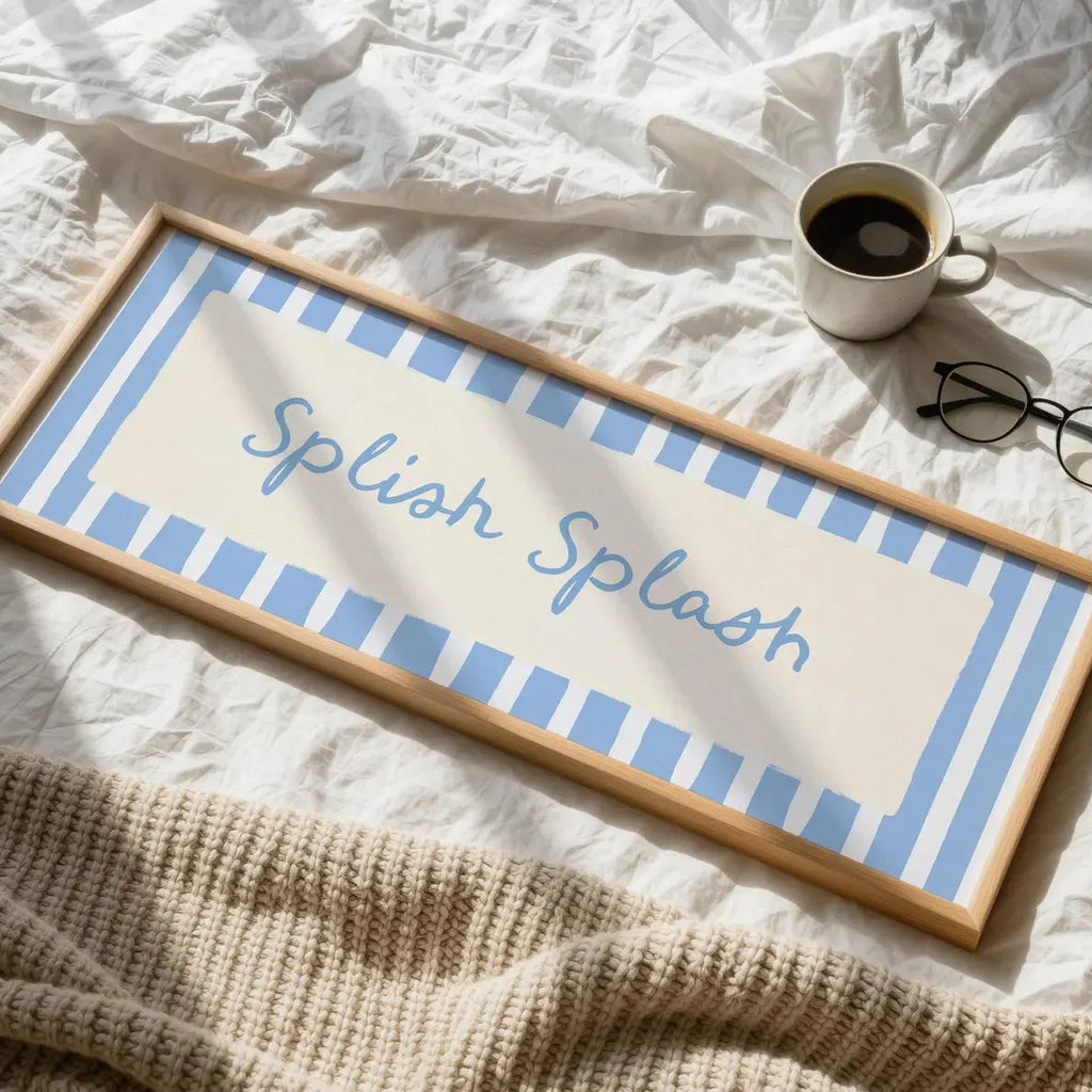 A rectangular framed sign with the words 'Splash Splash' in blue script, set on a rumpled white bed with a cup of coffee, eyeglasses, and a beige knit blanket nearby.