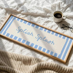 A rectangular framed sign with the words 'Splash Splash' in blue script, set on a rumpled white bed with a cup of coffee, eyeglasses, and a beige knit blanket nearby.
