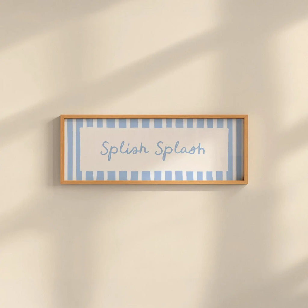 A rectangular framed wall sign with the words 'Splash Splash' in blue script, set against a light blue and white striped background with a light wood frame, mounted on a warm beige wall with soft diagonal shadows.