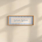 A rectangular framed wall sign with the words 'Splash Splash' in blue script, set against a light blue and white striped background with a light wood frame, mounted on a warm beige wall with soft diagonal shadows.