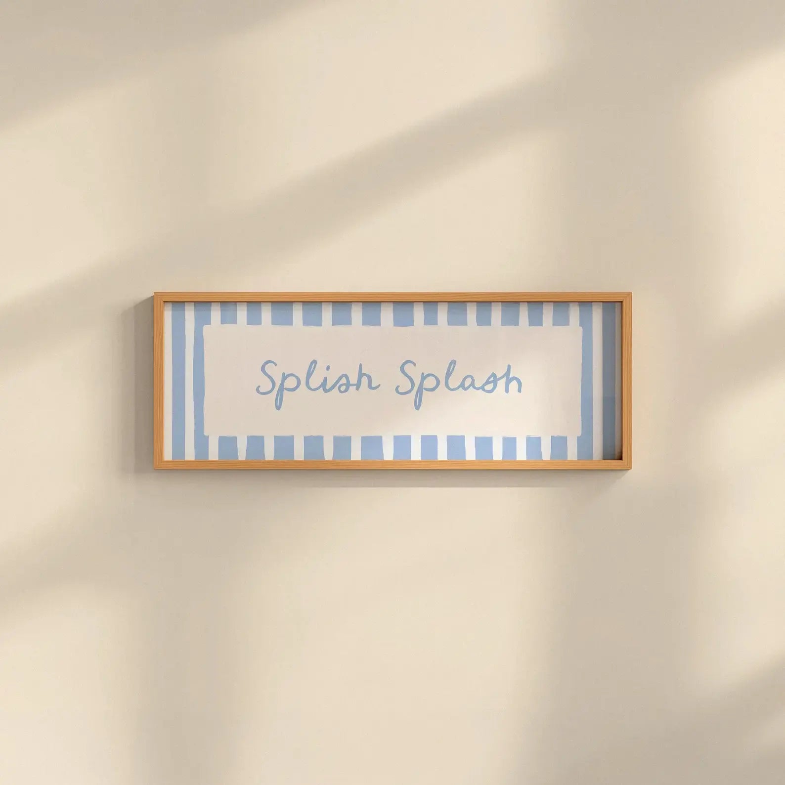 A rectangular framed wall sign with the words 'Splash Splash' in blue script, set against a light blue and white striped background with a light wood frame, mounted on a warm beige wall with soft diagonal shadows.