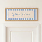 A framed decorative sign with the words 'Splash Splash' in blue script over a cream background and light blue vertical stripes, hung on a white wall above a white door.