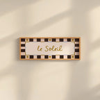 A rectangular wall sign with the French phrase "le Soleil" in gold cursive script, surrounded by a cream and black checkered border with small gold sun motifs. The sign is mounted on a warm beige wall with soft diagonal shadows.