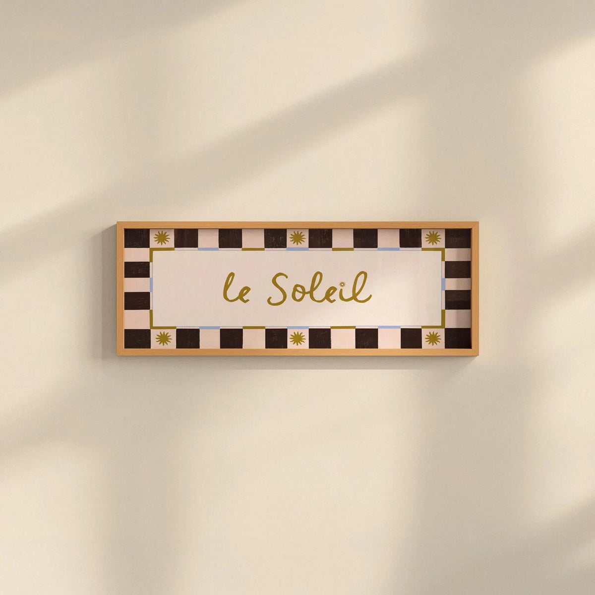 A rectangular wall sign with the French phrase "le Soleil" in gold cursive script, surrounded by a cream and black checkered border with small gold sun motifs. The sign is mounted on a warm beige wall with soft diagonal shadows.