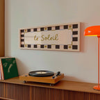 A framed wall art print with the text 'YOU ARE MY SUN, MY MOON AND ALL MY STARS' hangs on a light wall above a wooden credenza. The print has a black and cream checkered border with small gold decorative accents.