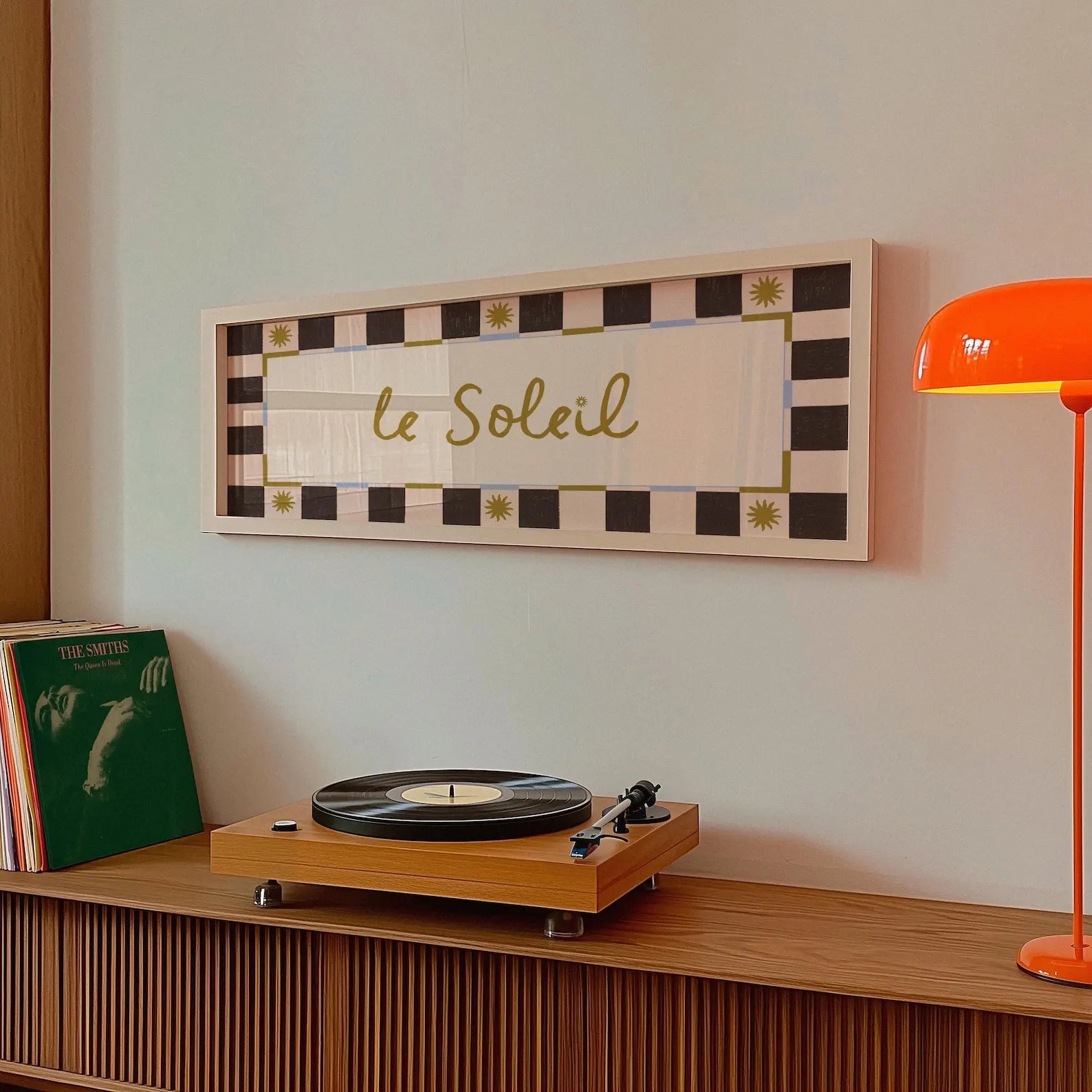 A framed wall art print with the text 'YOU ARE MY SUN, MY MOON AND ALL MY STARS' hangs on a light wall above a wooden credenza. The print has a black and cream checkered border with small gold decorative accents.