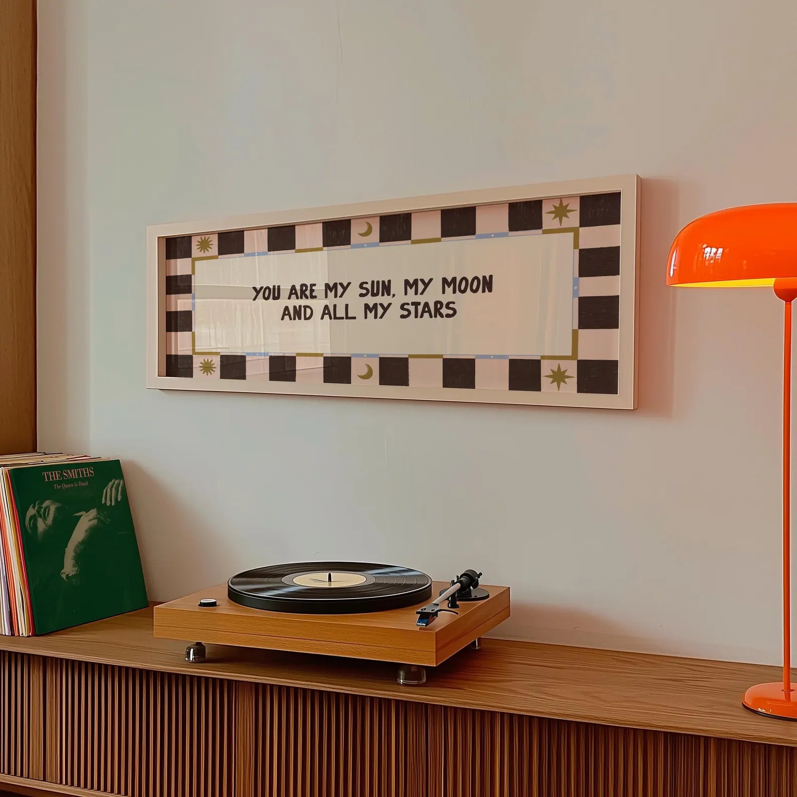 A framed wall art print with the text 'YOU ARE MY SUN, MY MOON AND ALL MY STARS' hangs on a light wall above a wooden credenza. The print has a black and cream checkered border with small gold decorative accents.