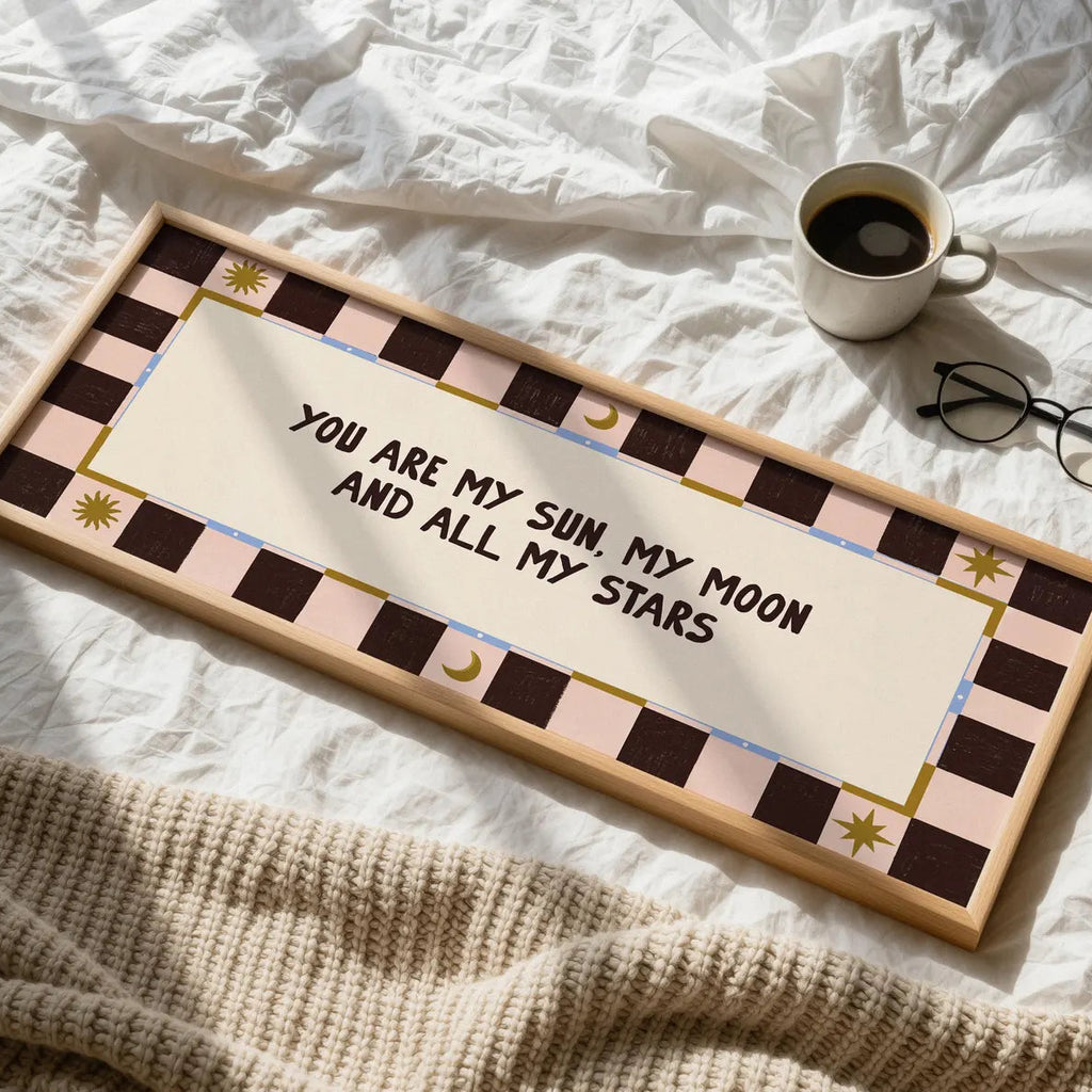 A rectangular wooden sign with a decorative pink, brown, and cream checker border and the phrase 'YOU ARE MY SUN, MY MOON AND ALL MY STARS' in black script, styled on a white bed with a coffee cup, glasses, and a knit blanket nearby.