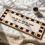 A rectangular wooden sign with a decorative pink, brown, and cream checker border and the phrase 'YOU ARE MY SUN, MY MOON AND ALL MY STARS' in black script, styled on a white bed with a coffee cup, glasses, and a knit blanket nearby.