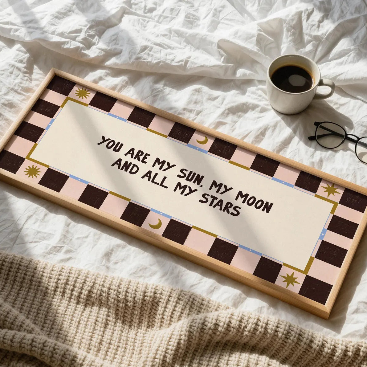 A rectangular wooden sign with a decorative pink, brown, and cream checker border and the phrase 'YOU ARE MY SUN, MY MOON AND ALL MY STARS' in black script, styled on a white bed with a coffee cup, glasses, and a knit blanket nearby.