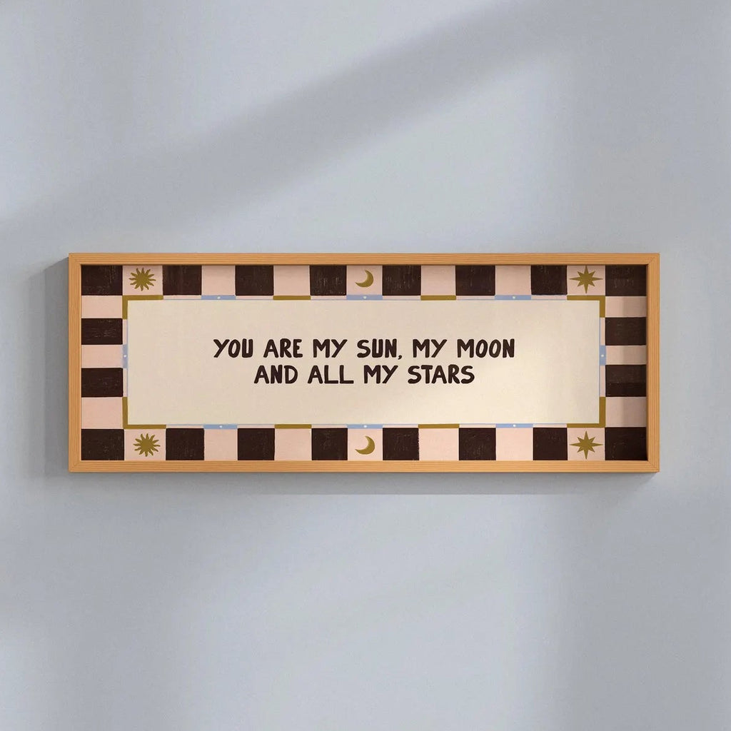 A rectangular wall sign with a warm beige center panel and black text reading 'YOU ARE MY SUN, MY MOON AND ALL MY STARS,' surrounded by a brown and beige checkered border with small gold decorative motifs, mounted on a light gray wall.