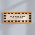 A rectangular wall sign with a warm beige center panel and black text reading 'YOU ARE MY SUN, MY MOON AND ALL MY STARS,' surrounded by a brown and beige checkered border with small gold decorative motifs, mounted on a light gray wall.