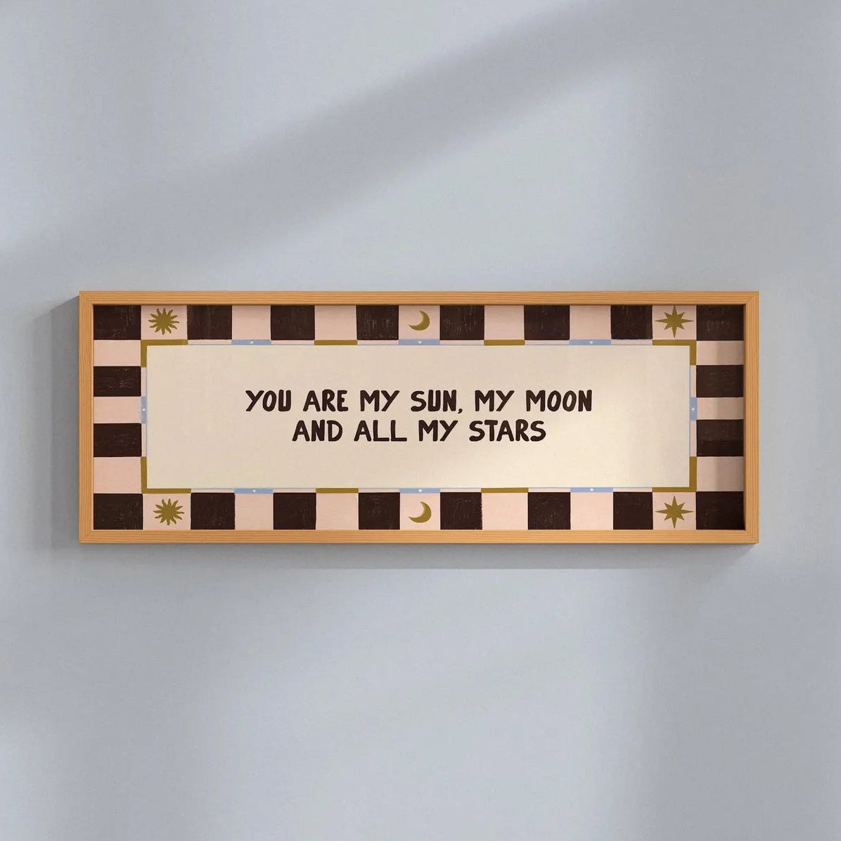 A rectangular wall sign with a warm beige center panel and black text reading 'YOU ARE MY SUN, MY MOON AND ALL MY STARS,' surrounded by a brown and beige checkered border with small gold decorative motifs, mounted on a light gray wall.