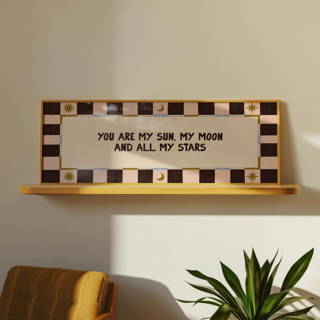 A rectangular wall sign with the phrase 'YOU ARE MY SUN, MY MOON AND ALL MY STARS' in black lettering, mounted on a light wood shelf against a beige wall. The sign has a decorative border with black and beige checkered stripes and small sun and moon motifs.