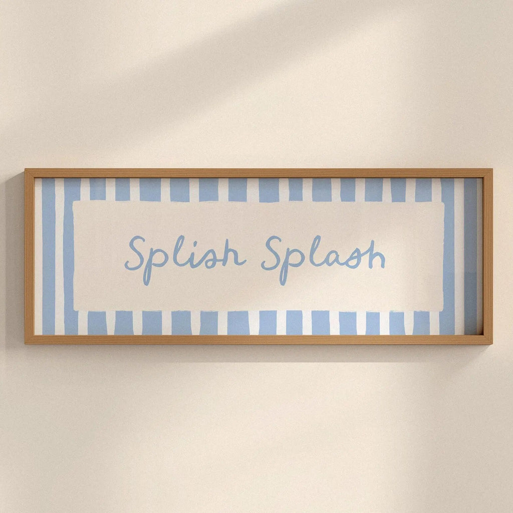 A framed wall art print with the words 'Splash Splash' in light blue script, set against a background of light blue and white vertical stripes. The frame is a natural light wood, mounted on a warm beige wall with soft shadows.