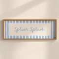 A framed wall art print with the words 'Splash Splash' in light blue script, set against a background of light blue and white vertical stripes. The frame is a natural light wood, mounted on a warm beige wall with soft shadows.