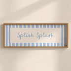 A framed wall art print with the words 'Splash Splash' in light blue script, set against a background of light blue and white vertical stripes. The frame is a natural light wood, mounted on a warm beige wall with soft shadows.