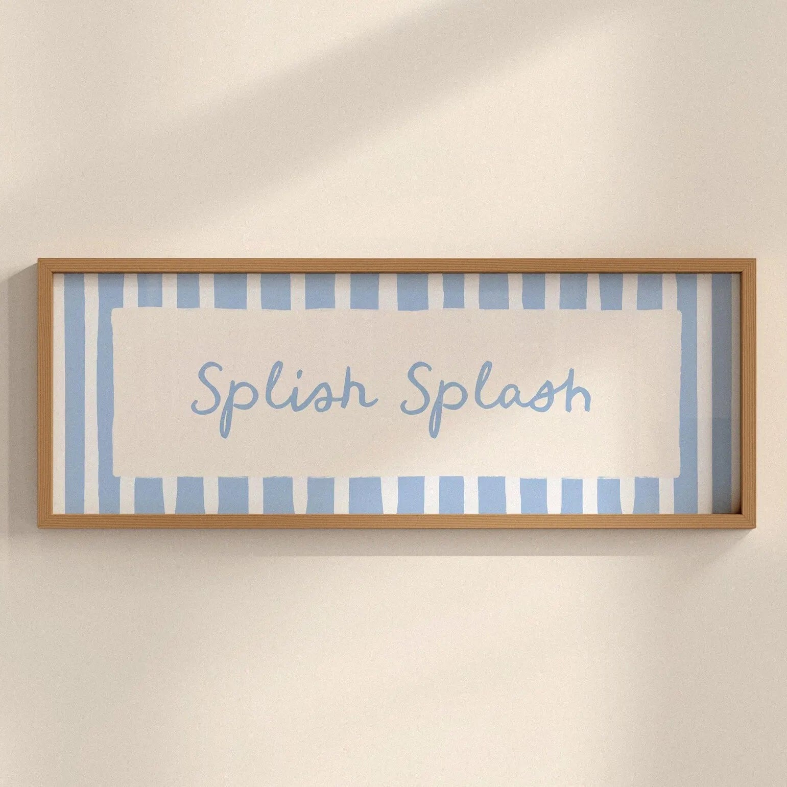 A framed wall art print with the words 'Splash Splash' in light blue script, set against a background of light blue and white vertical stripes. The frame is a natural light wood, mounted on a warm beige wall with soft shadows.