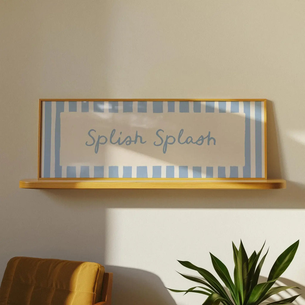 A framed wall art print with the words 'Splash Splash' in blue script over a light blue and white striped background, displayed on a wooden floating shelf in a styled interior setting with a plant and chair partially visible.