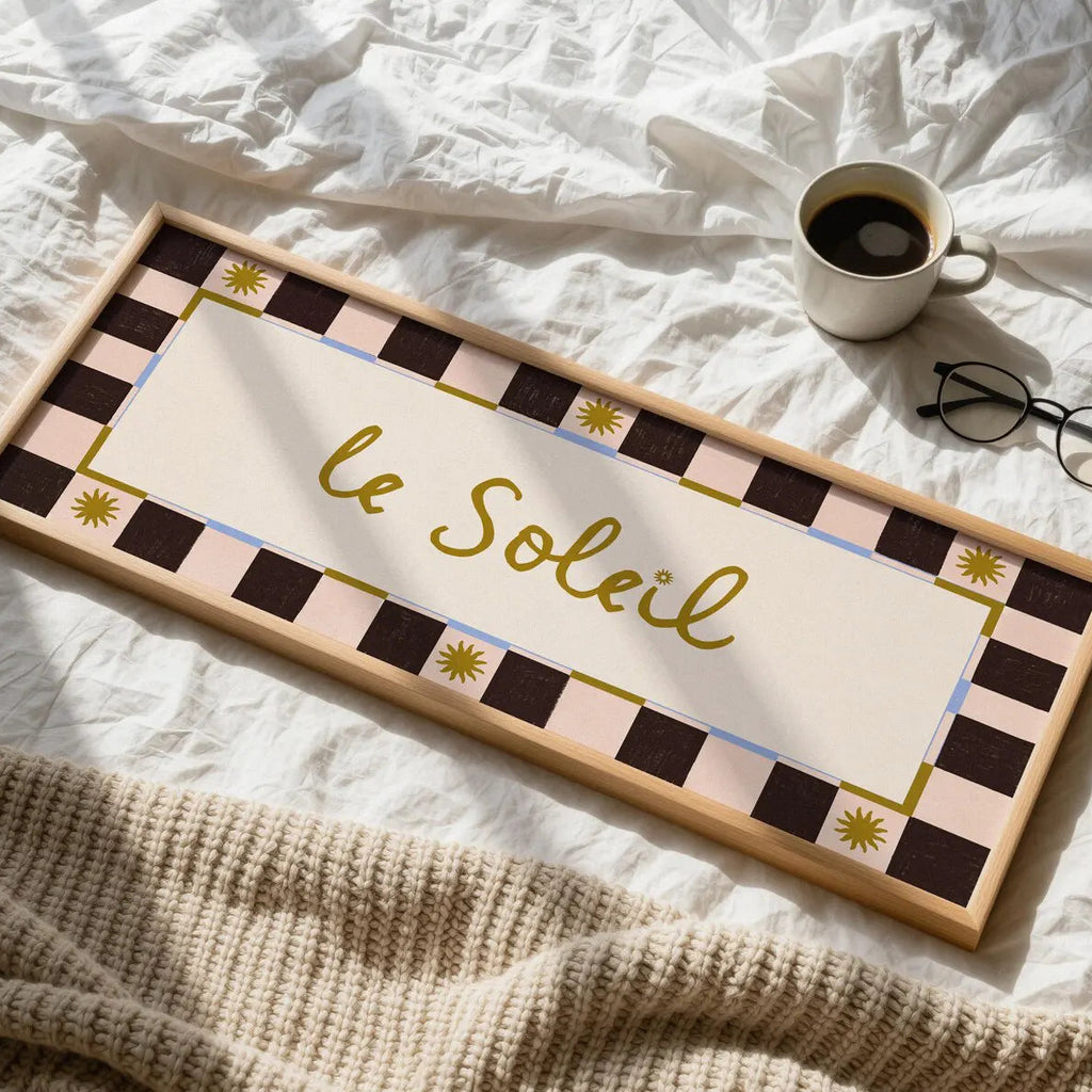 A rectangular wooden sign with the French phrase “le Soleil” in gold script, surrounded by a cream center panel and a dark brown and pale pink checkered border with small gold sun motifs. The sign is styled on white bed sheets with a beige knit blanket, a cup of coffee, and a pair of eyeglasses nearby.