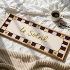 A rectangular wooden sign with the French phrase “le Soleil” in gold script, surrounded by a cream center panel and a dark brown and pale pink checkered border with small gold sun motifs. The sign is styled on white bed sheets with a beige knit blanket, a cup of coffee, and a pair of eyeglasses nearby.