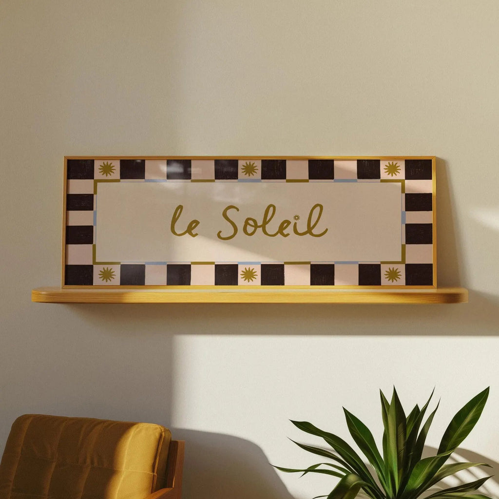 A rectangular decorative wall sign with the words "le Soleil" in gold script, mounted on a light wood shelf against a warm beige wall. The sign has a cream center panel and a border of alternating black and pale pink checkered squares with small gold sun motifs.
