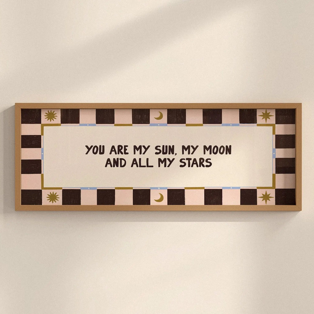 A framed wall art print with the text 'YOU ARE MY SUN, MY MOON AND ALL MY STARS' in dark brown lettering on a cream center panel. The frame and border feature a checkered pattern in dark brown, light pink, and beige, with small gold decorative sun and moon motifs.