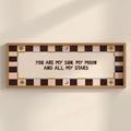 A framed wall art print with the text 'YOU ARE MY SUN, MY MOON AND ALL MY STARS' in dark brown lettering on a cream center panel. The frame and border feature a checkered pattern in dark brown, light pink, and beige, with small gold decorative sun and moon motifs.