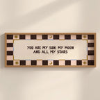 A framed wall art print with the text 'YOU ARE MY SUN, MY MOON AND ALL MY STARS' in dark brown lettering on a cream center panel. The frame and border feature a checkered pattern in dark brown, light pink, and beige, with small gold decorative sun and moon motifs.