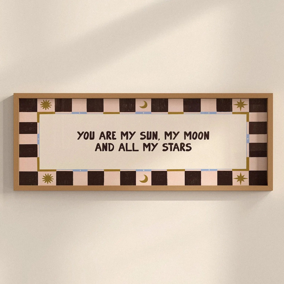 A framed wall art print with the text 'YOU ARE MY SUN, MY MOON AND ALL MY STARS' in dark brown lettering on a cream center panel. The frame and border feature a checkered pattern in dark brown, light pink, and beige, with small gold decorative sun and moon motifs.