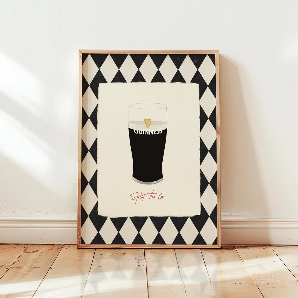 A framed art print leaning against a white wall on a light wood floor. The print features a black and white harlequin diamond border surrounding an illustration of a pint of Guinness with the word 'GUINNESS' and the phrase 'Split the G' in red script below.