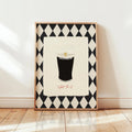 A framed art print leaning against a white wall on a light wood floor. The print features a black and white harlequin diamond border surrounding an illustration of a pint of Guinness with the word 'GUINNESS' and the phrase 'Split the G' in red script below.