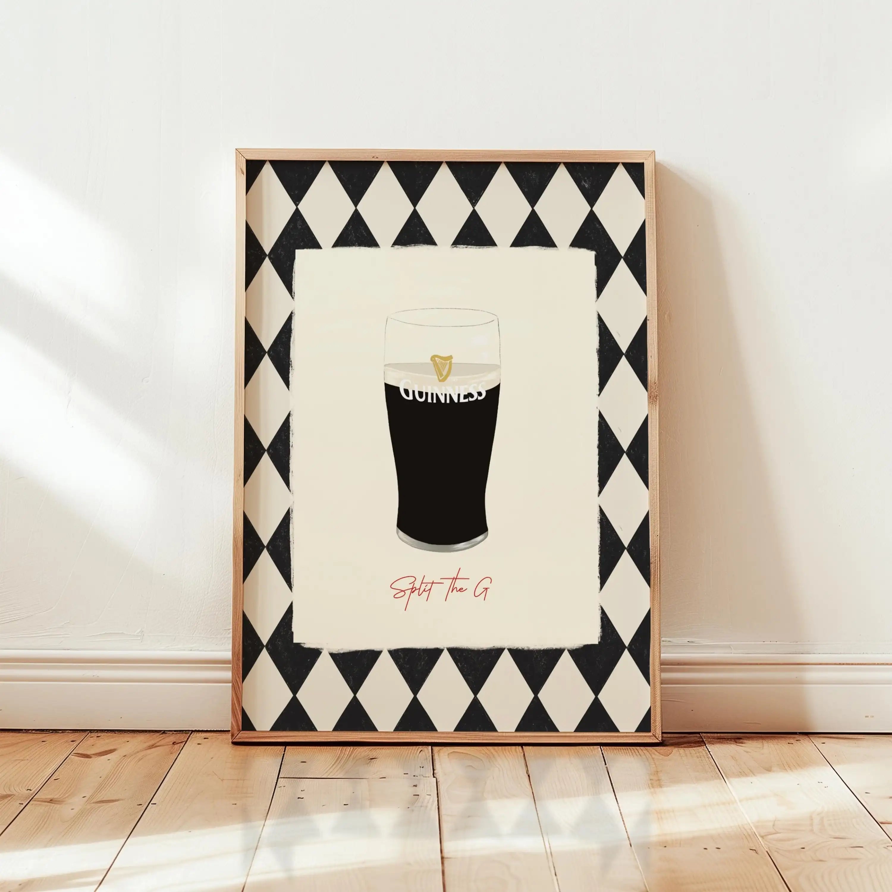 A framed art print leaning against a white wall on a light wood floor. The print features a black and white harlequin diamond border surrounding an illustration of a pint of Guinness with the word 'GUINNESS' and the phrase 'Split the G' in red script below.