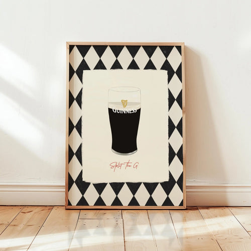 A framed art print leaning against a white wall on a light wood floor. The print features a black and white harlequin diamond border surrounding an illustration of a pint of Guinness with the word 'GUINNESS' and the phrase 'Split the G' in red script below.